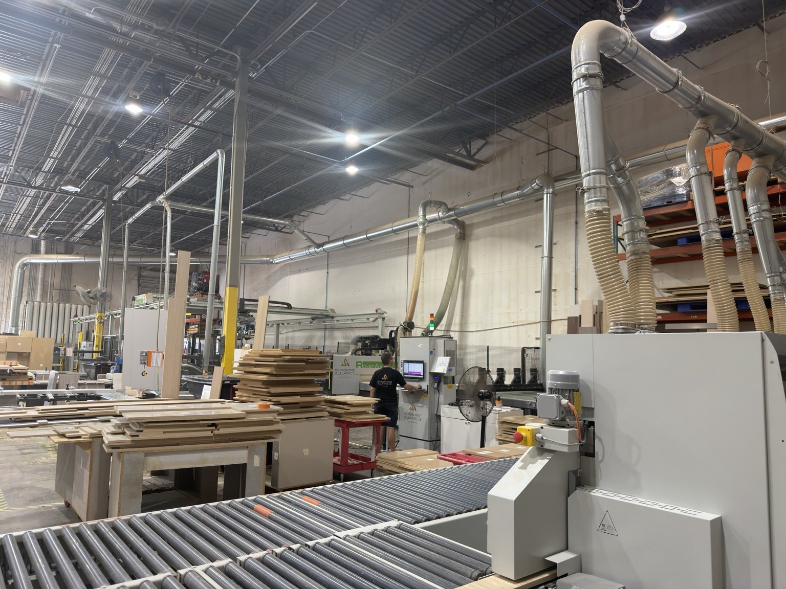 Wide view down the cabinet factory floor with overhead dust collection, roller conveyor and stacked cut parts | Sunshine Alliance, Deerfield Beach FL